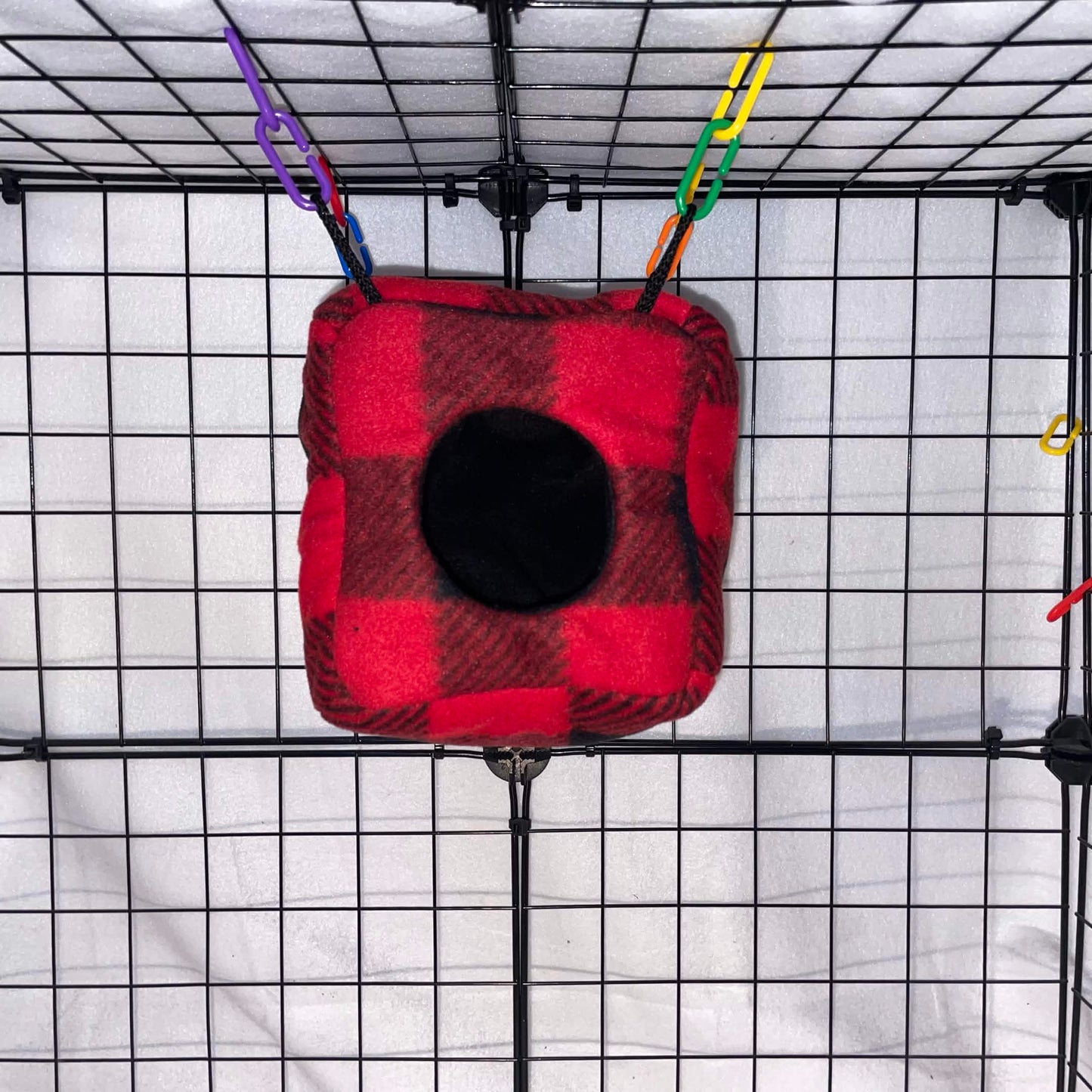 Red buffalo plaid sugar glider cube with black fleece interior and small opening hanging inside a wire display cage.