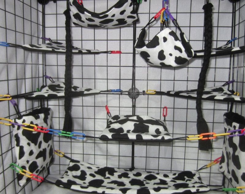 17-piece sugar glider cage set featuring cow print fleece fabric, including hammocks, pouches, and accessories arranged inside a metal cage for small pet comfort and enrichment.