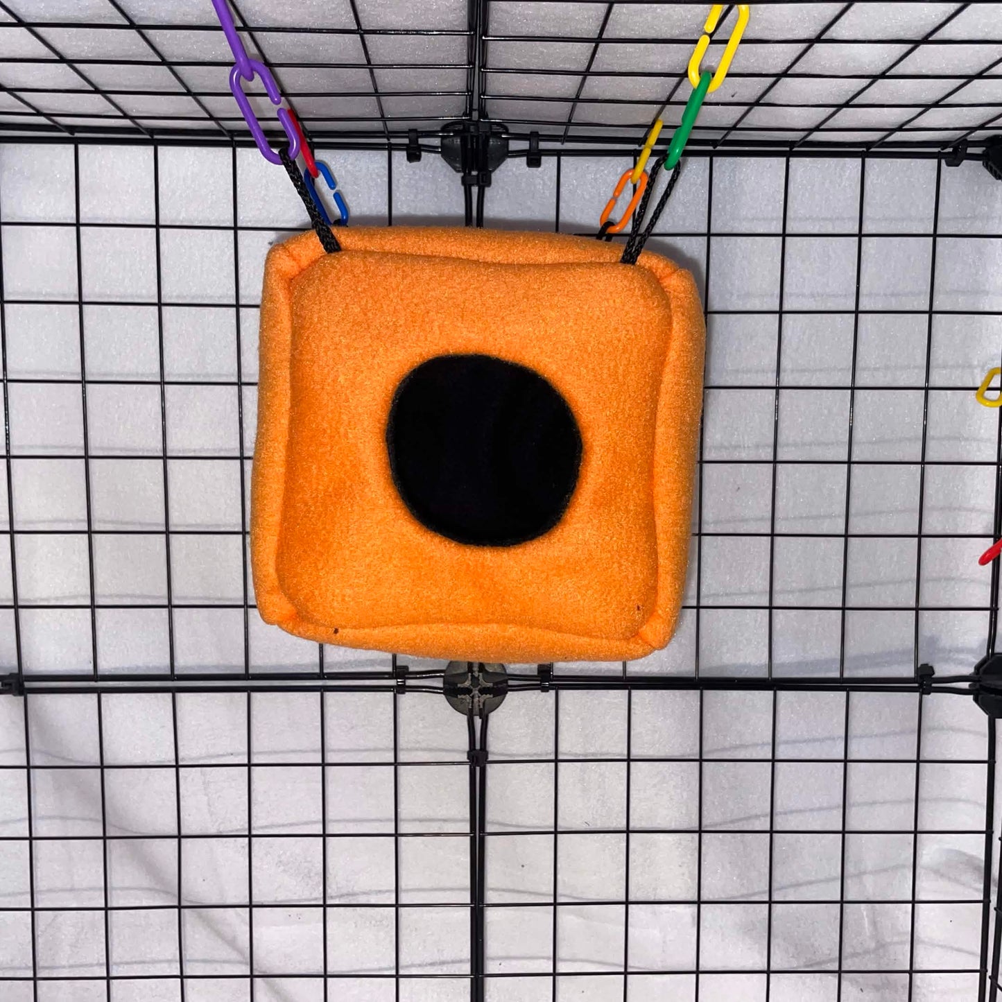 Orange sugar glider fleece cube hideout with black opening hanging in a wire cage.