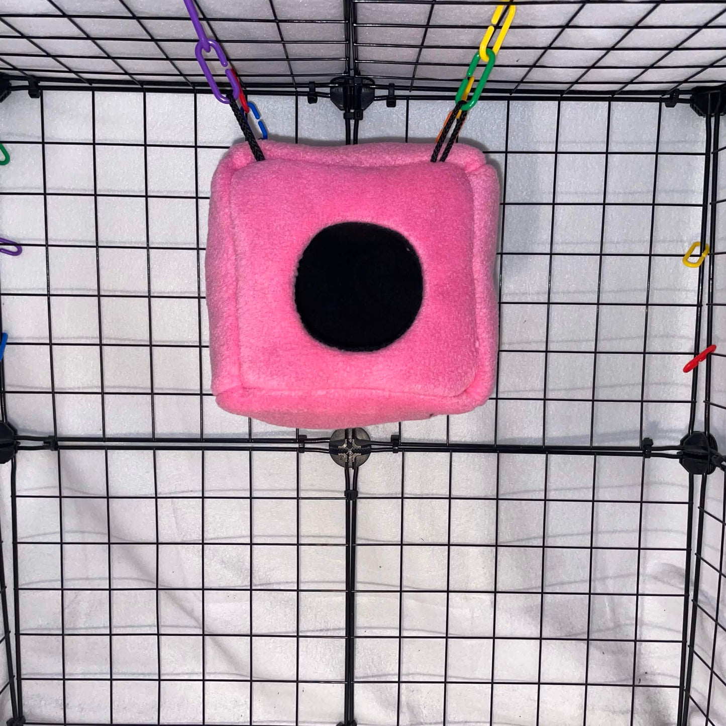 Pink sugar glider fleece cube hideout with black opening hanging in a wire cage.