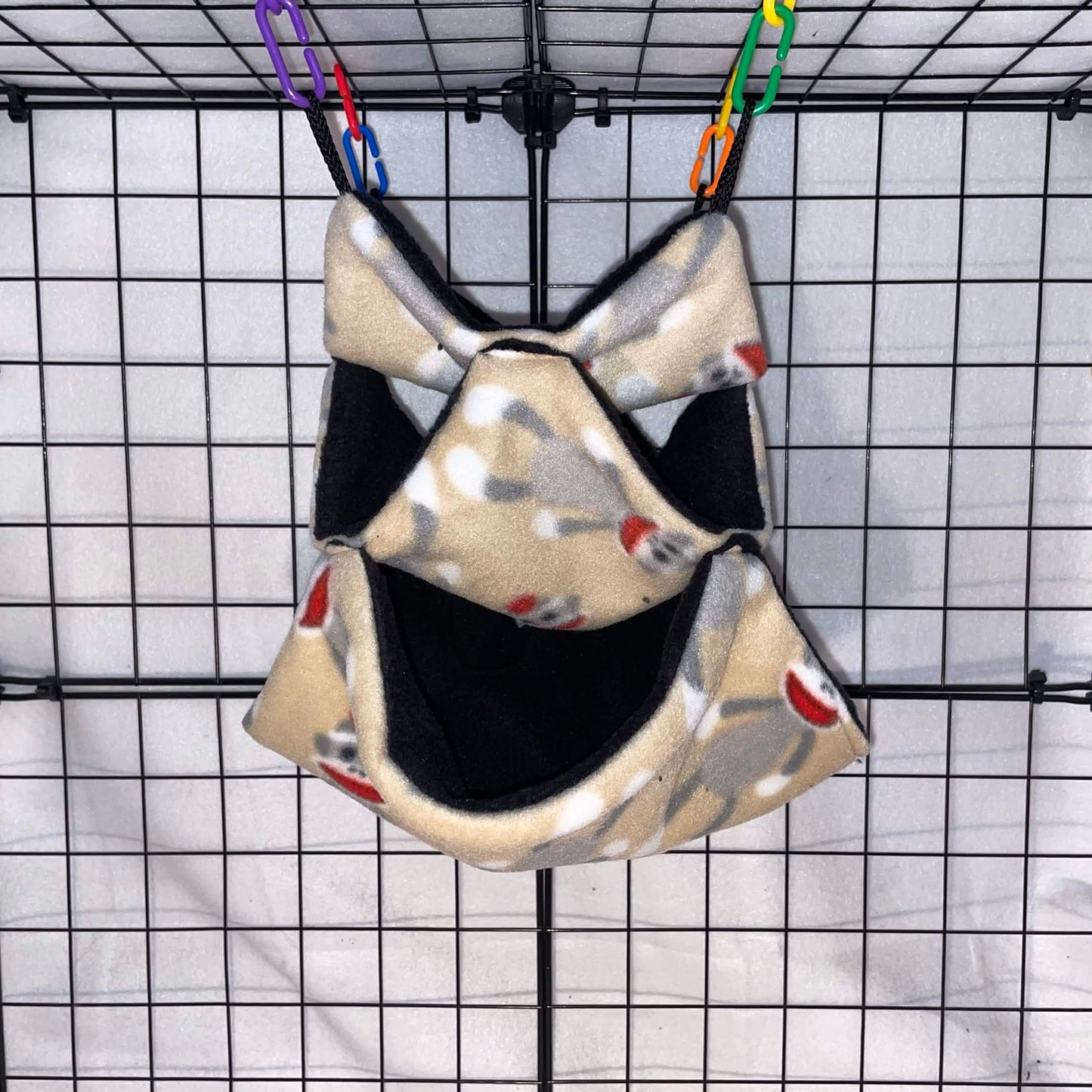 Sock monkey print sugar glider 3-tier honeycomb hammock with black interior hanging in a wire cage.